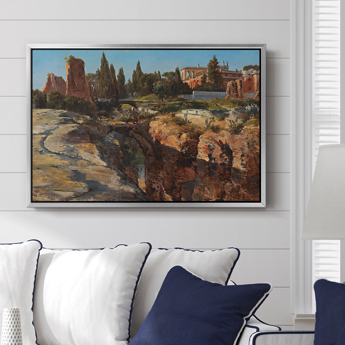Palatine in Rome, c. 1921 by Edward Theodore Compton displayed above a sofa in a living room in a silver floater frame, featuring an architectural scene of ancient structures in sunlit environment.
