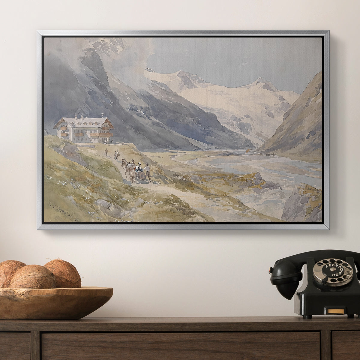 Shepherds (Pasterze), c. 1921 by Edward Theodore Compton in a silver floater framed canvas displayed above a console table in a hallway, featuring a natural landscape with human presence among mountains.