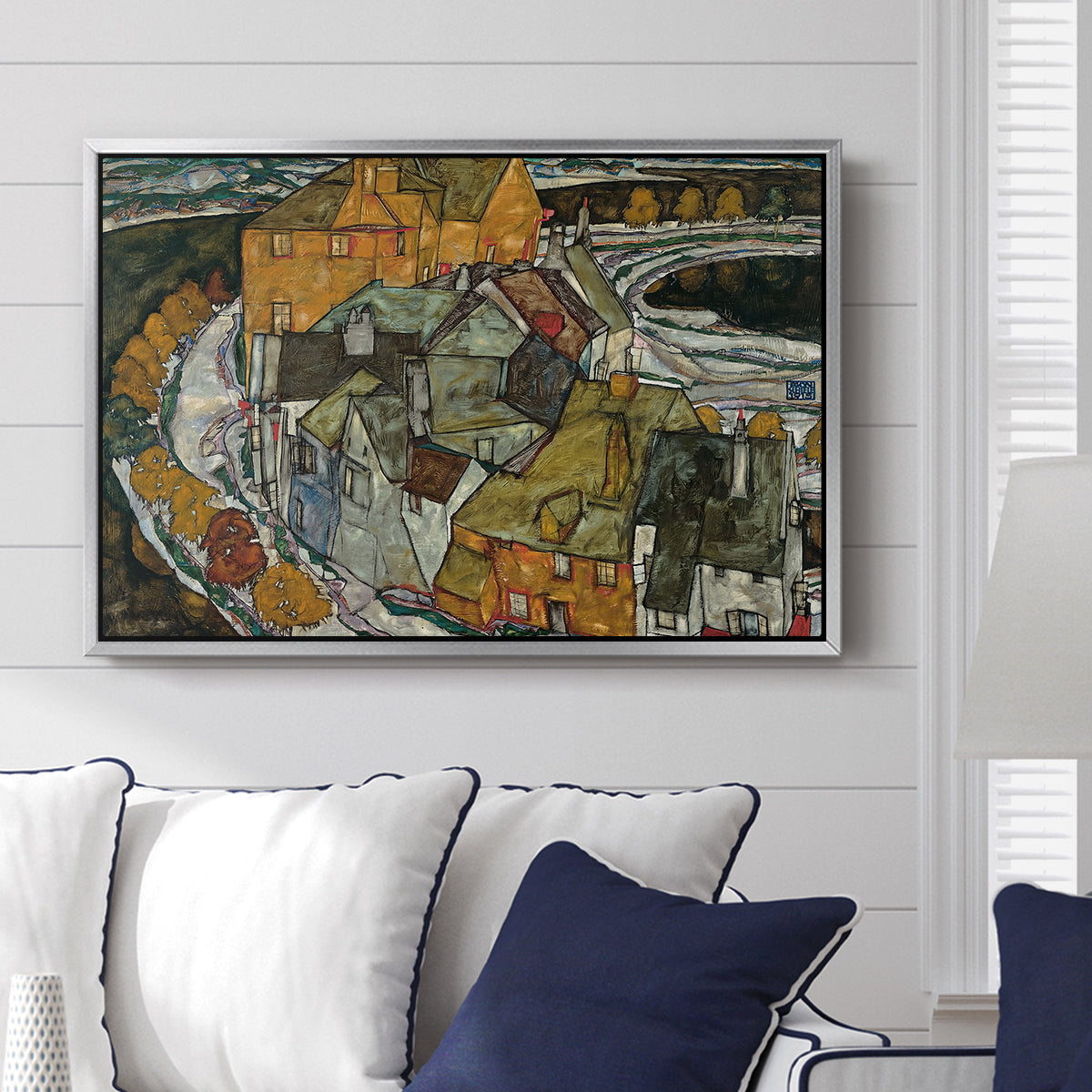 Crescent of Houses II (Island Town), 1915 by Egon Schiele displayed above a sofa in a living room in a silver floater frame, featuring a stylized townscape with clustered buildings and bold outlines.