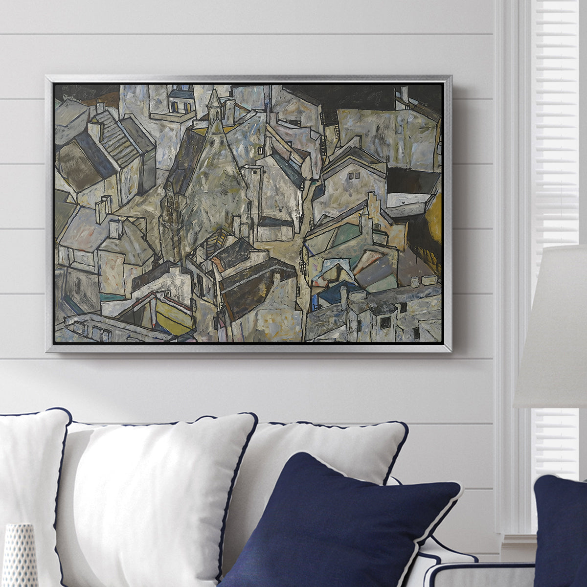 Mödling II, 1918 by Egon Schiele displayed above a sofa in a living room in a silver floater frame, featuring an expressionist cityscape with tightly packed buildings and bold lines.