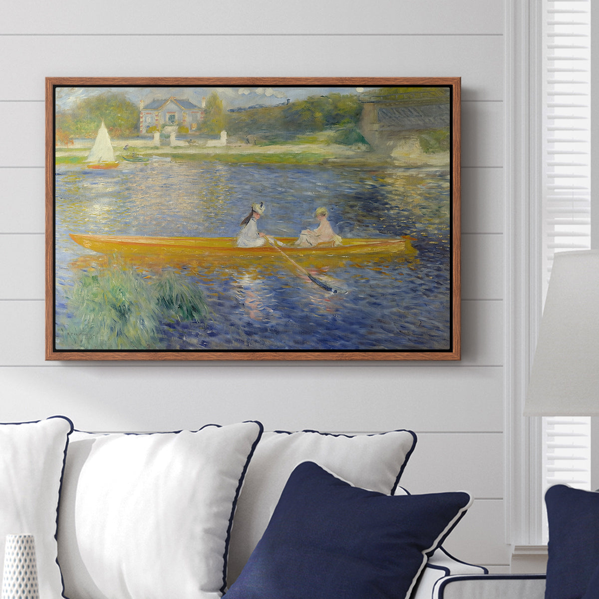 The Skiff (La Yole), 1875 by Pierre-Auguste Renoir displayed above a sofa in a living room in a walnut floater frame, featuring an impressionist boating scene with people relaxing in a narrow skiff.