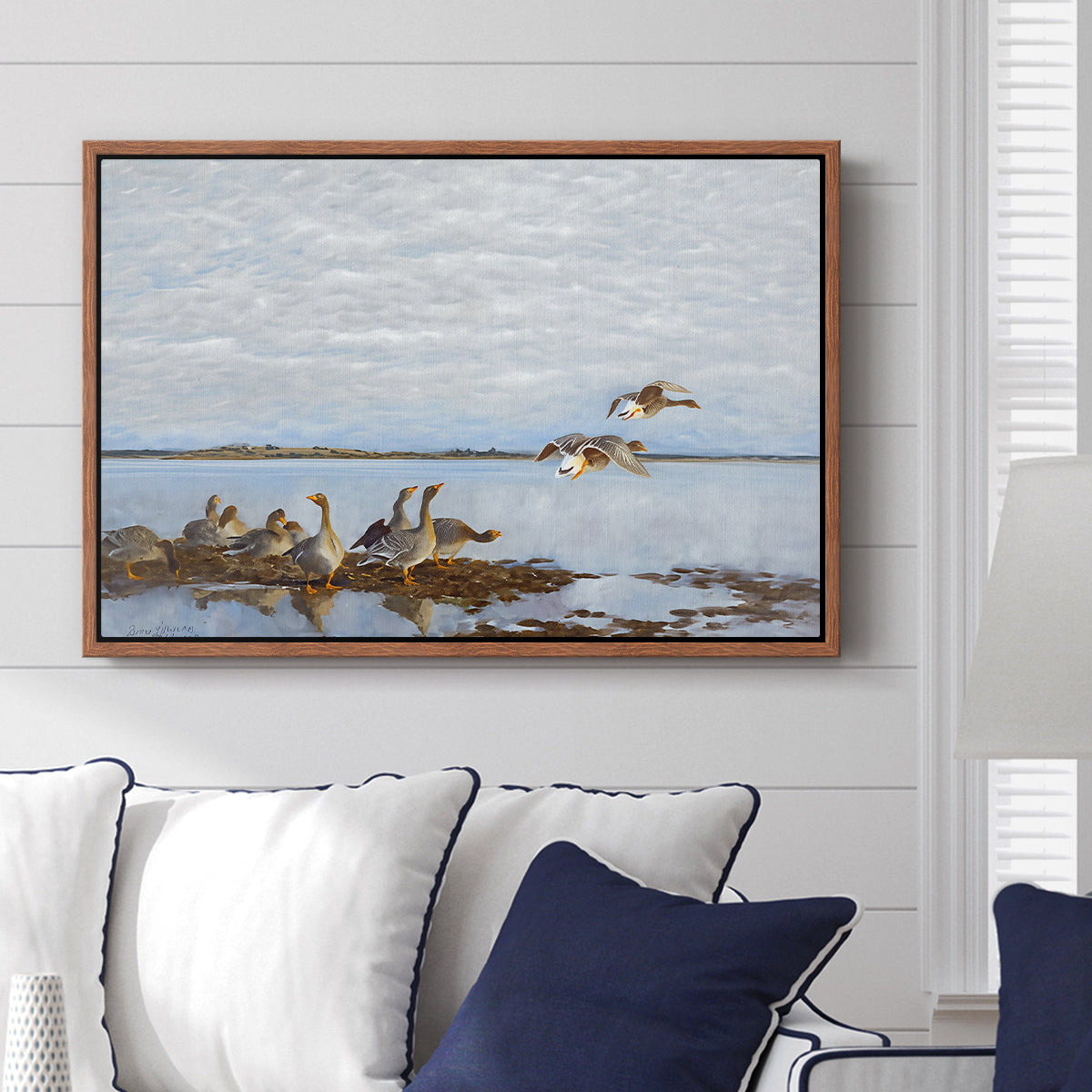 Greylag Geese (Grågäss), 1922 by Bruno Liljefors displayed above a sofa in a living room in a walnut floater frame, featuring birds standing and resting along shoreline with soft grass and water.