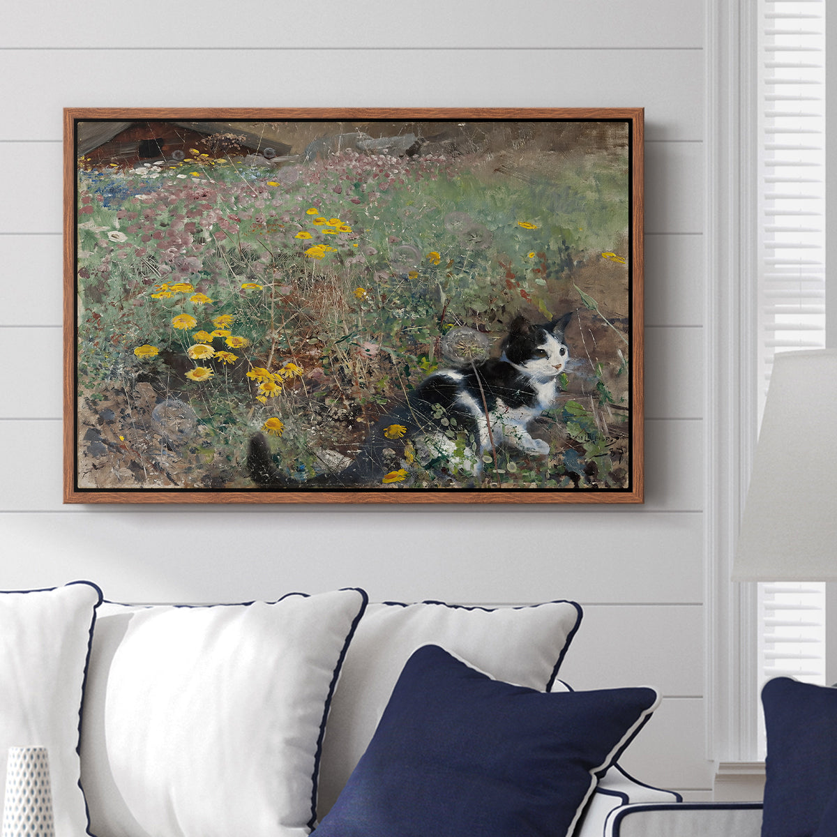 Cat on a Flowery Meadow, 1887 by Bruno Liljefors displayed above a sofa in a living room in a walnut floater frame, featuring a small cat resting among colorful flowers in grassy field.