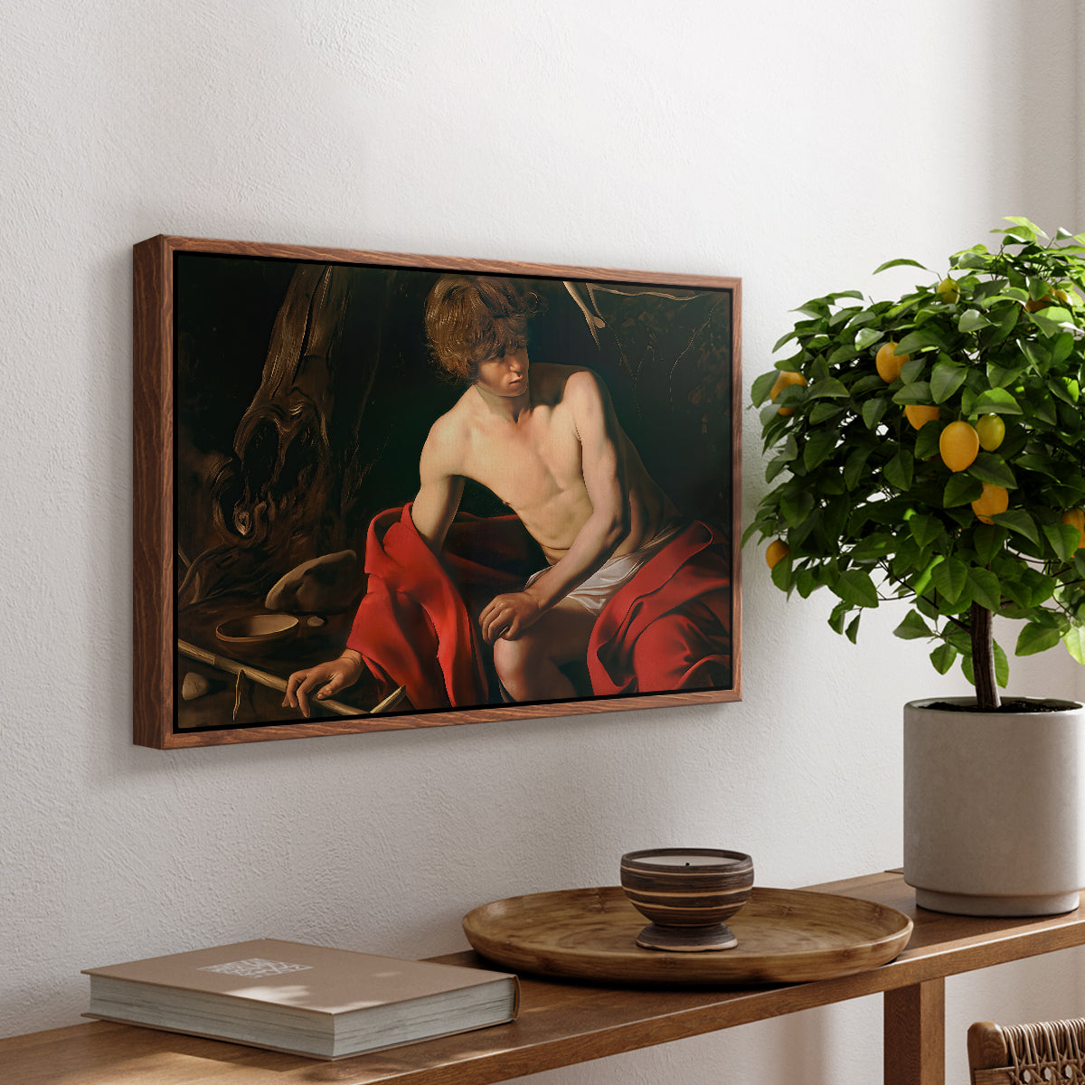Saint John the Baptist in the Wilderness, c. 1603-1604 by Caravaggio, presented in a walnut floater framed canvas featuring a young Saint John seated in wilderness with staff and red drapery, displayed above a hallway console table.