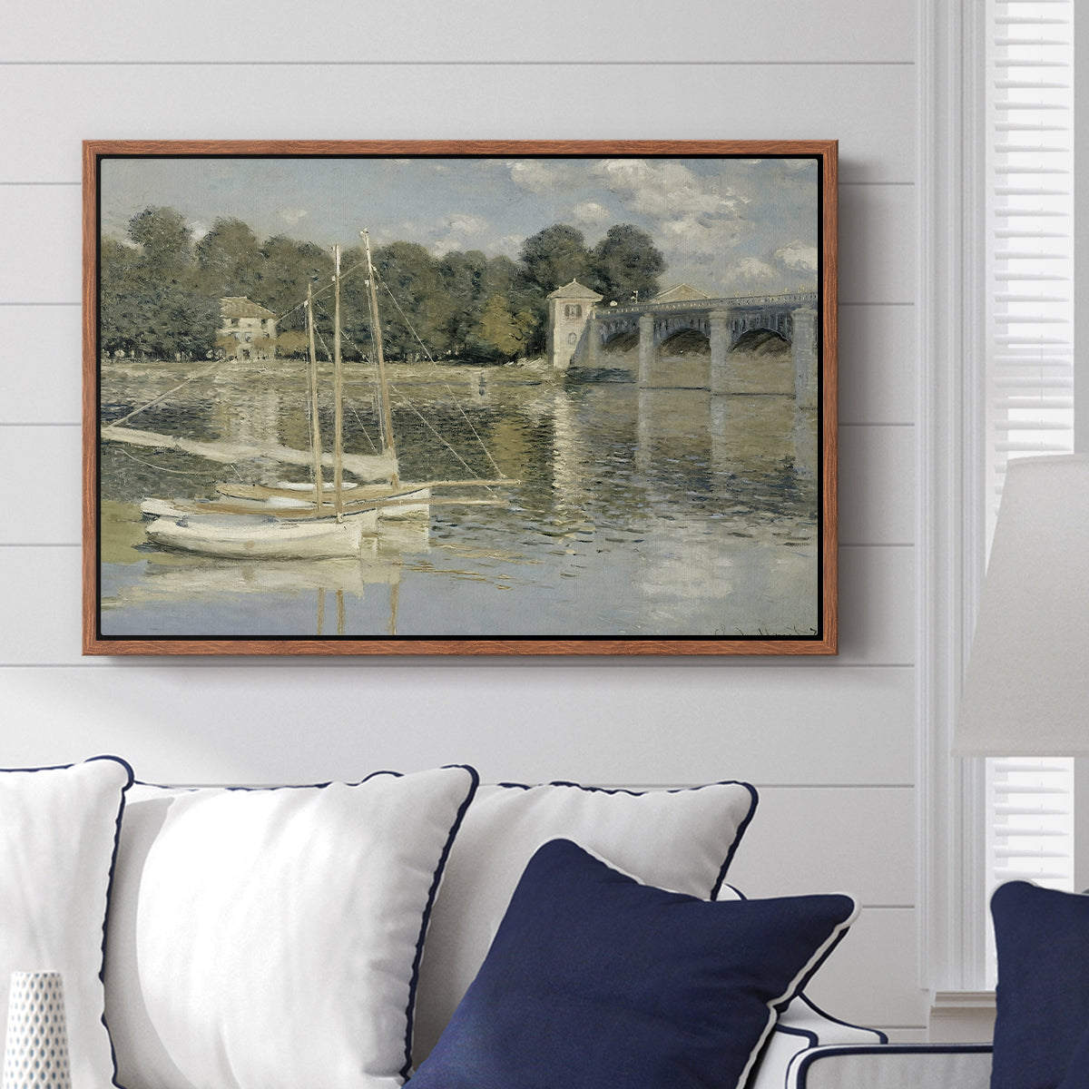 The Bridge at Argenteuil, 1874 by Claude Monet displayed above a sofa in a living room in a walnut floater frame, featuring an impressionist river scene with boats passing beneath bridge.