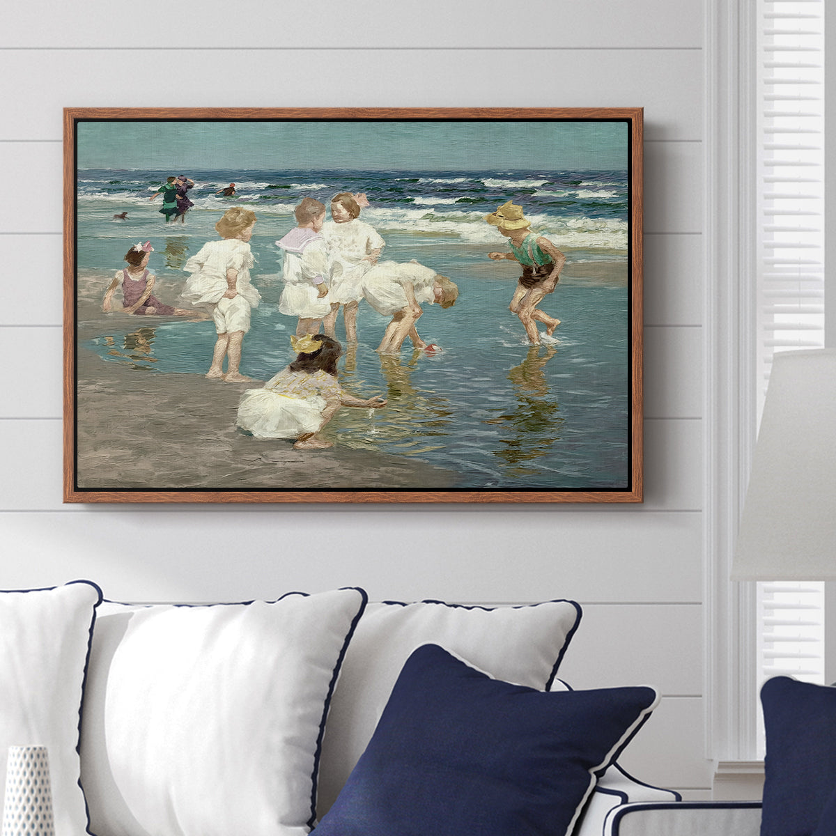 A Holiday, c. 1915 by Edward Henry Potthast displayed above a sofa in a living room in a walnut floater frame, featuring a beach scene with figures bathing and standing in waves under bright sky.
