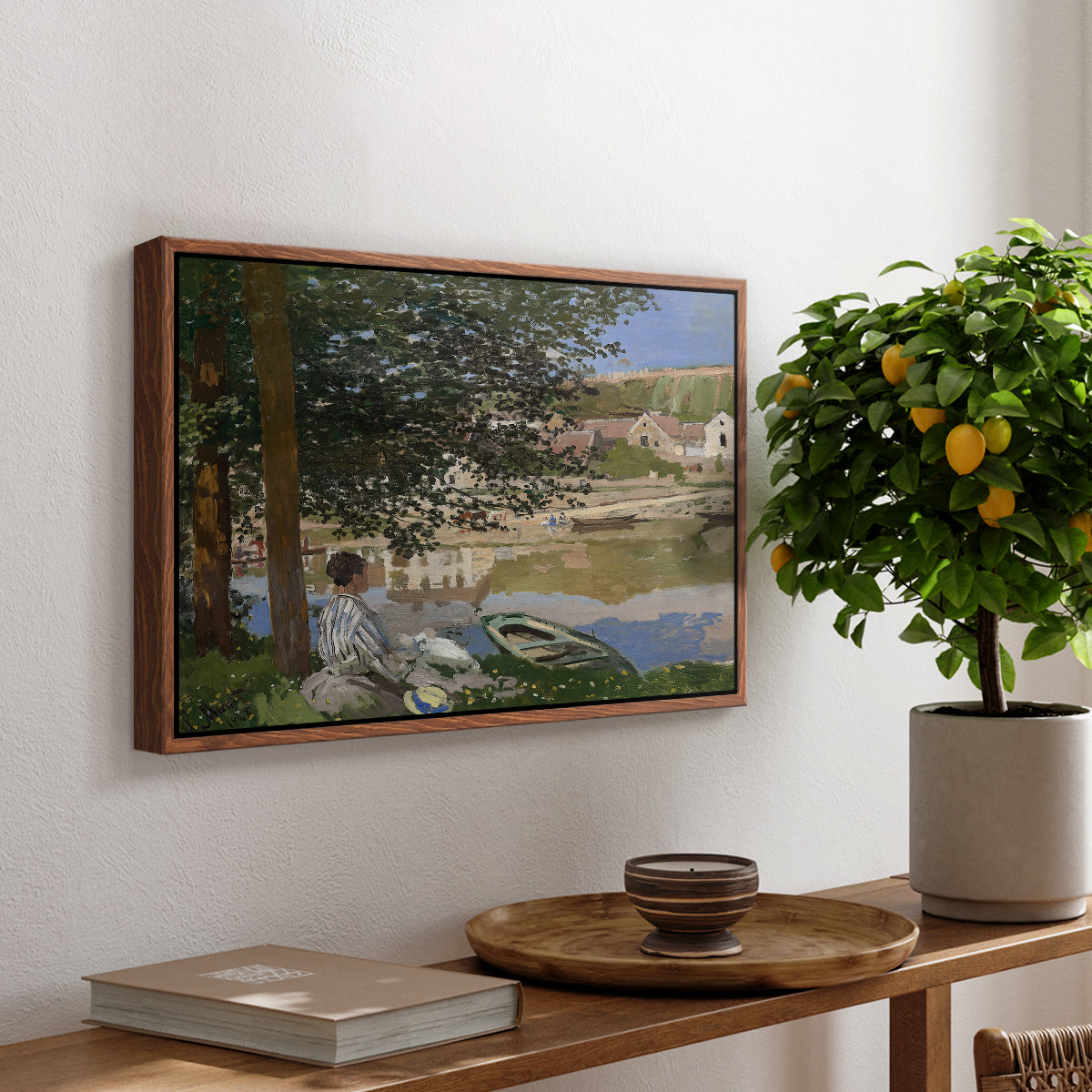 On the Bank of the Seine, Bennecourt, 1868 by Claude Monet, presented in a walnut floater framed canvas featuring a woman and child seated along riverbank beneath trees in soft daylight, displayed above a hallway console table.