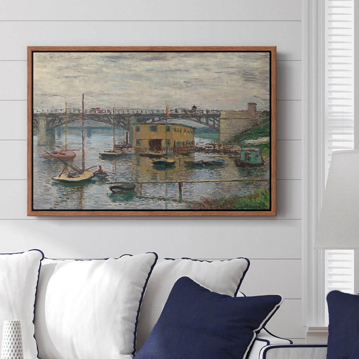 Bridge at Argenteuil on a Gray Day, c. 1876 by Claude Monet displayed above a sofa in a living room in a walnut floater frame, featuring an impressionist riverscape with overcast atmosphere and soft tones.