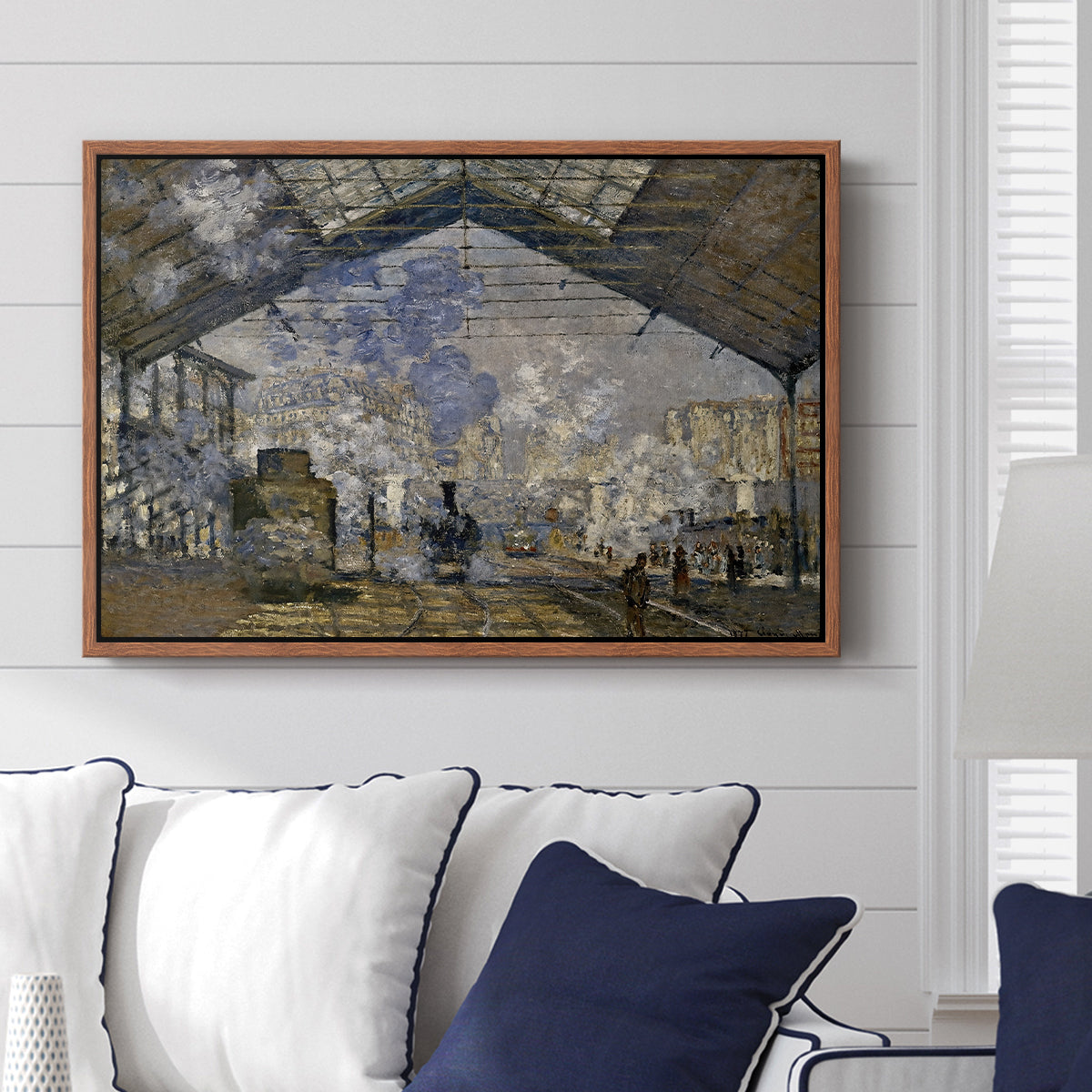 The Gare Saint-Lazare, 1877 by Claude Monet displayed above a sofa in a living room in a walnut floater frame, featuring an impressionist scene of trains and smoke inside busy railway station.