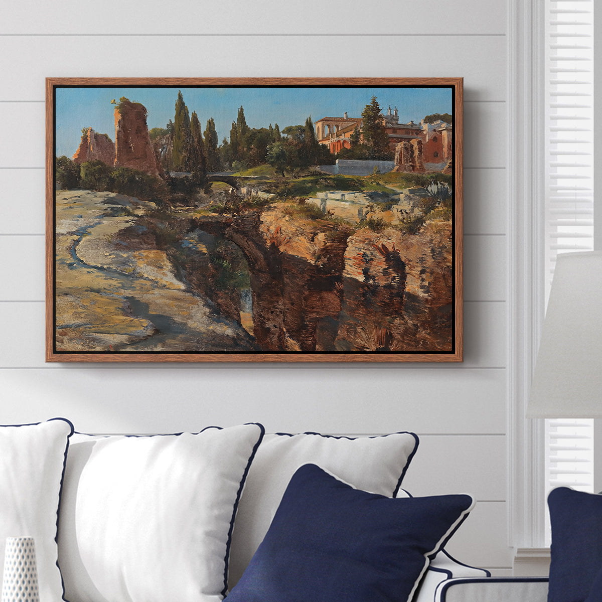 Palatine in Rome, c. 1921 by Edward Theodore Compton displayed above a sofa in a living room in a walnut floater frame, featuring an architectural scene of ancient structures in sunlit environment.