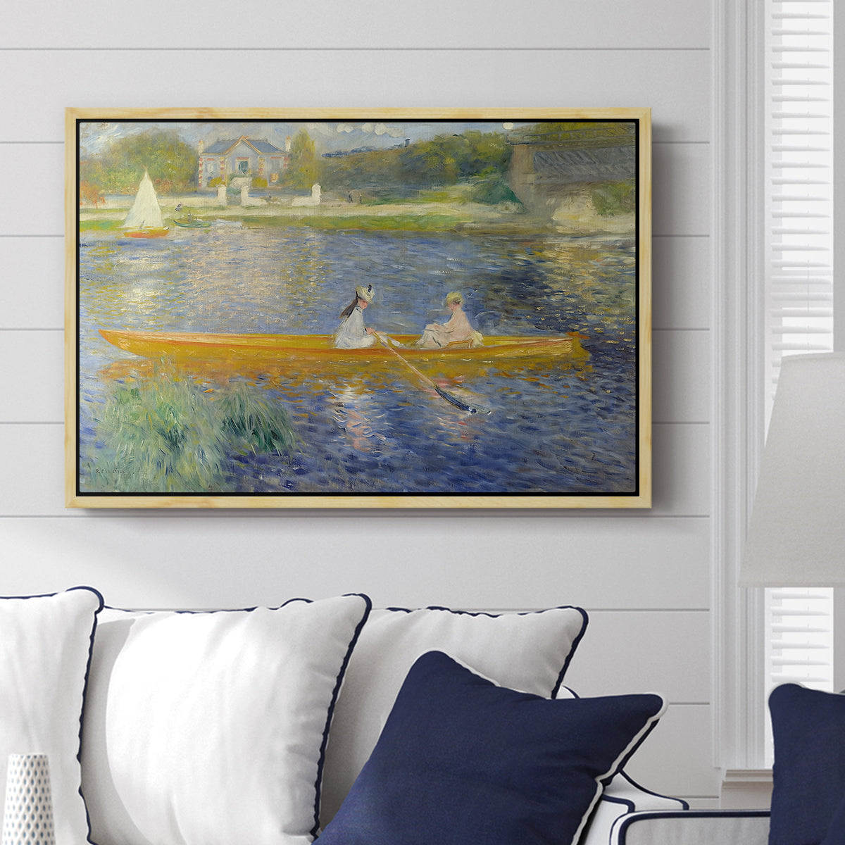 The Skiff (La Yole), 1875 by Pierre-Auguste Renoir displayed above a sofa in a living room in a natural wood floater frame, featuring an impressionist boating scene with people relaxing in a narrow skiff.