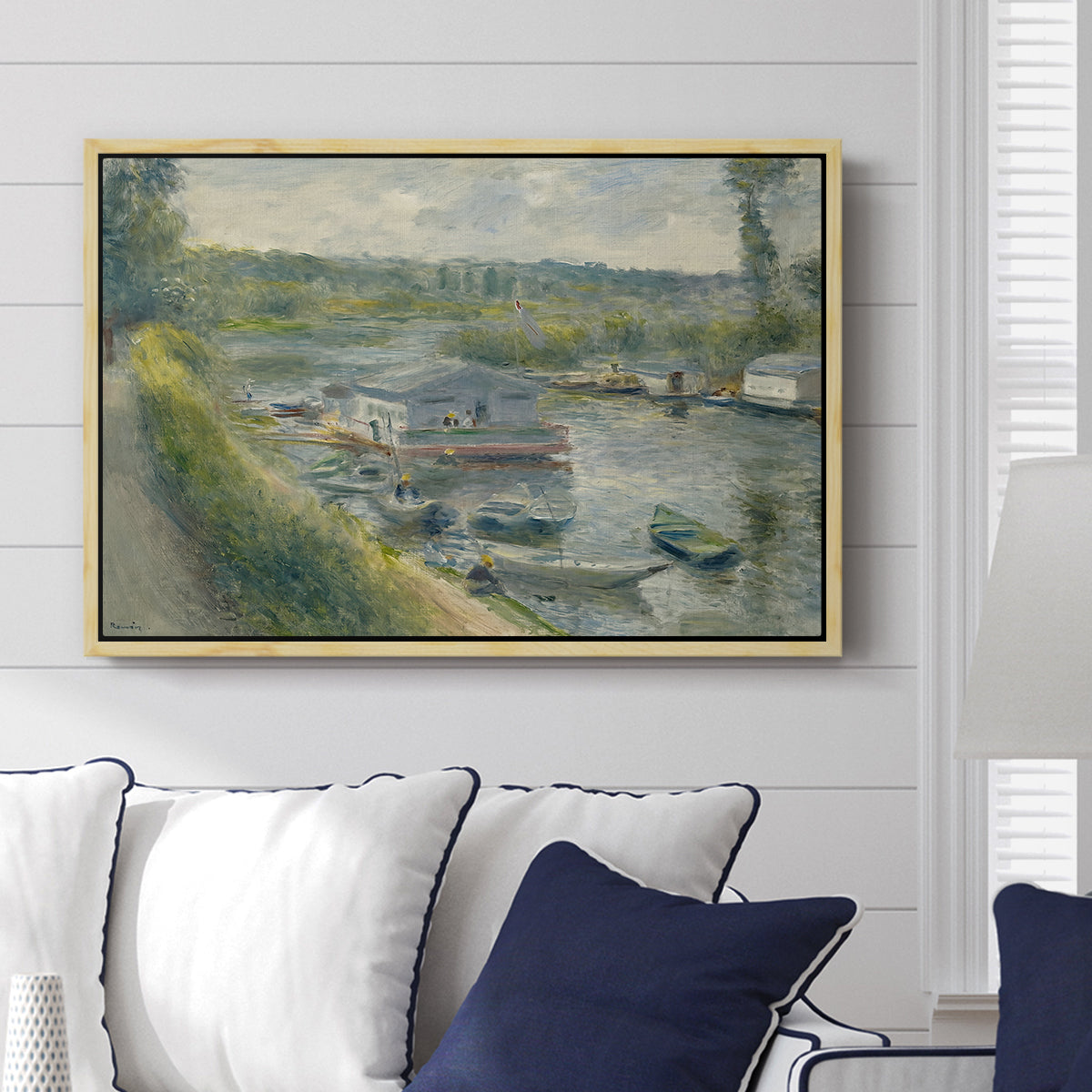 Wash-House Boat at Bas-Meudon, c. 1874 by Pierre-Auguste Renoir displayed above a sofa in a living room in a natural wood floater frame, featuring a riverside scene with moored boat used for washing clothes.