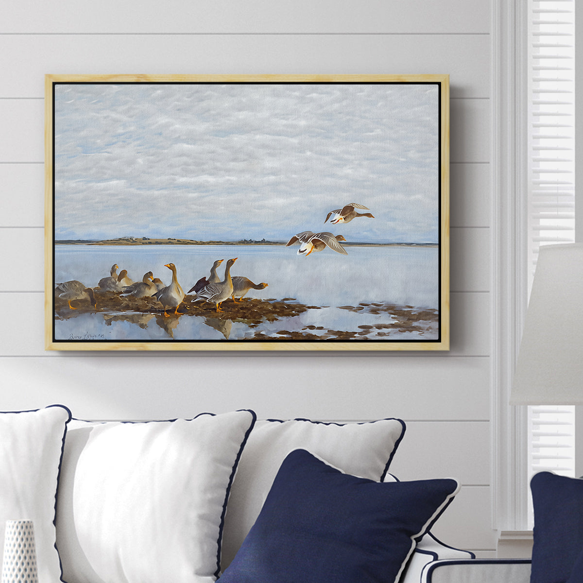 Greylag Geese (Grågäss), 1922 by Bruno Liljefors displayed above a sofa in a living room in a natural wood floater frame, featuring birds standing and resting along shoreline with soft grass and water.
