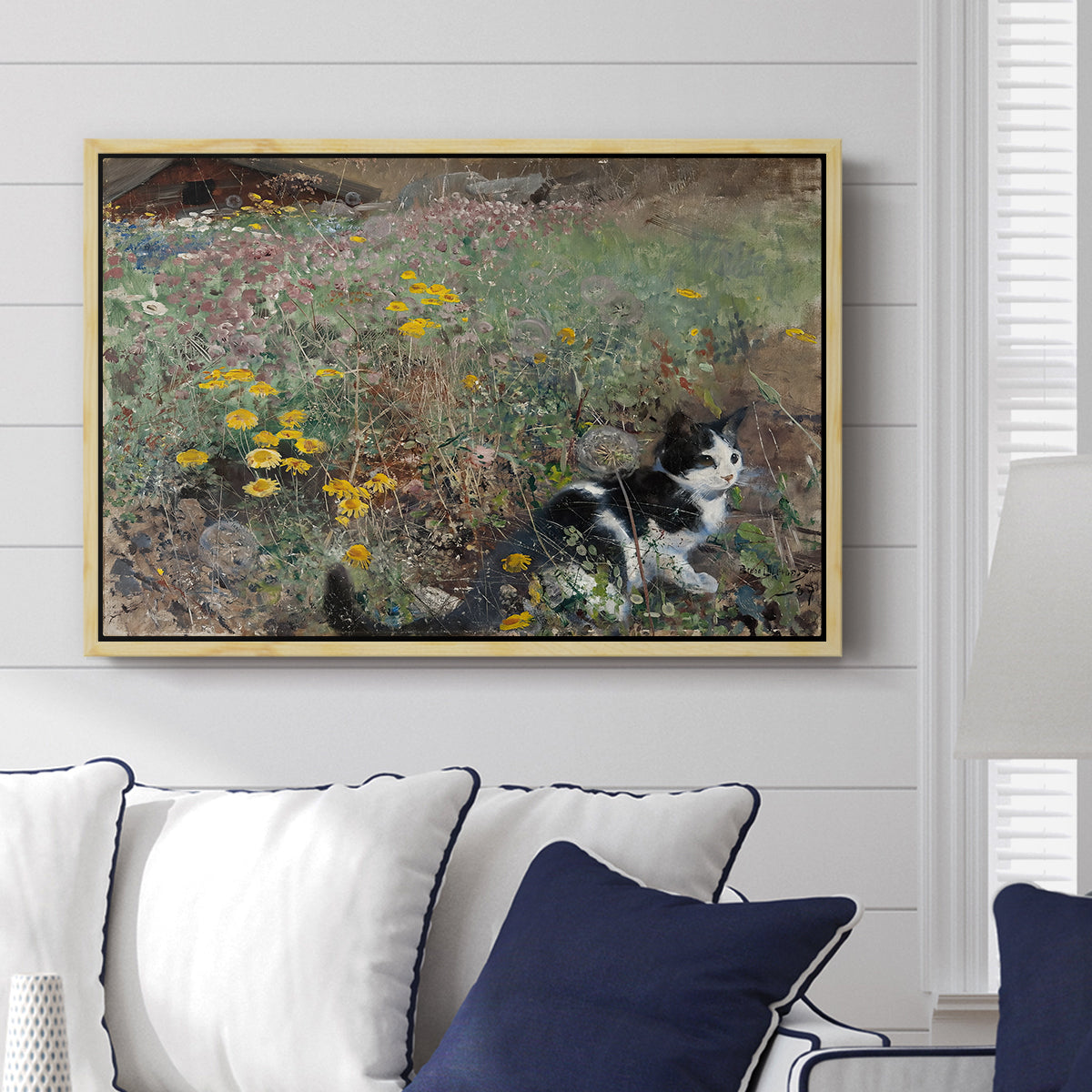Cat on a Flowery Meadow, 1887 by Bruno Liljefors displayed above a sofa in a living room in a natural wood floater frame, featuring a small cat resting among colorful flowers in grassy field.