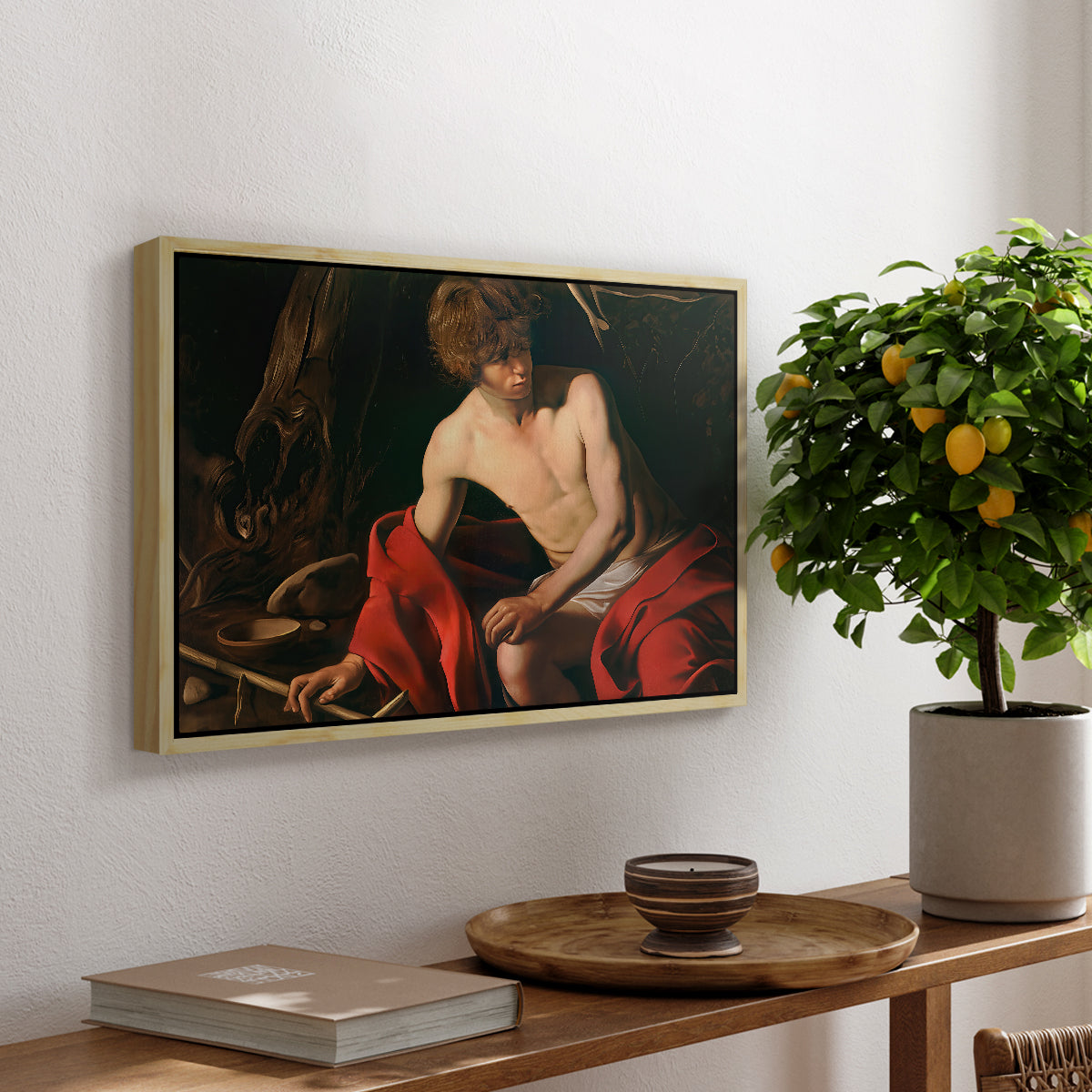 Saint John the Baptist in the Wilderness, c. 1603-1604 by Caravaggio, presented in a natural wood floater framed canvas featuring a young Saint John seated in wilderness with staff and red drapery, displayed above a hallway console table.