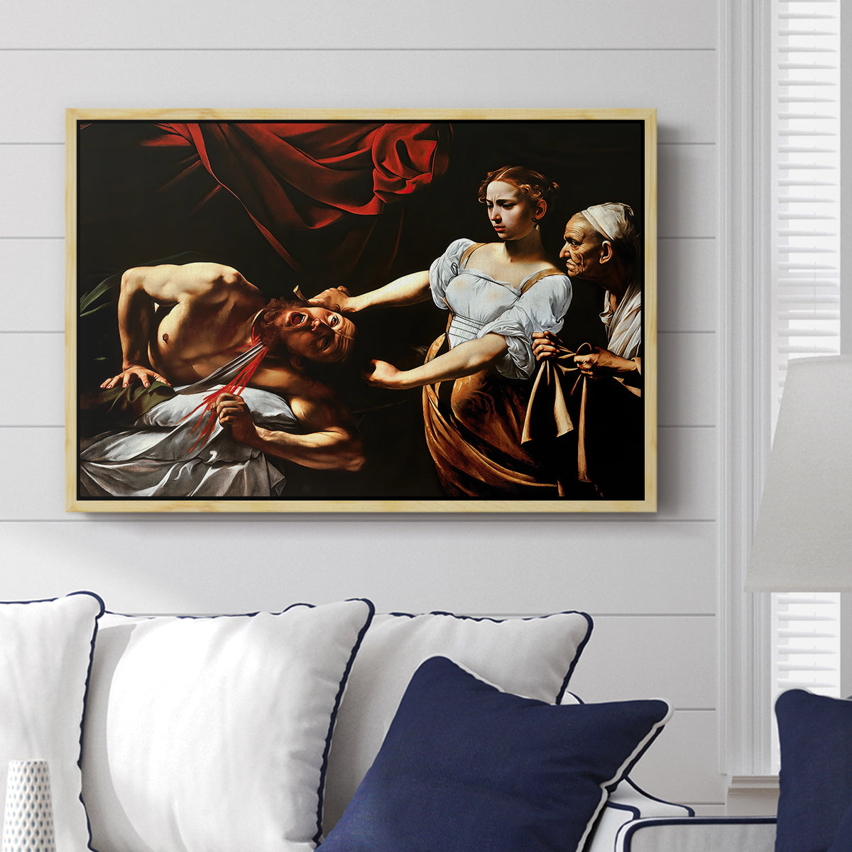 Judith Beheading Holofernes, c. 1599 by Caravaggio displayed above a sofa in a living room in a natural wood floater frame, featuring a dramatic biblical scene of woman beheading man in bed.
