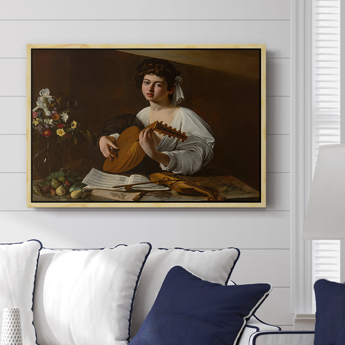 The Lute Player (Hermitage Version), c. 1596 by Caravaggio displayed above a sofa in a living room in a natural wood floater frame, featuring a portrait of lute player surrounded by still life arrangement.