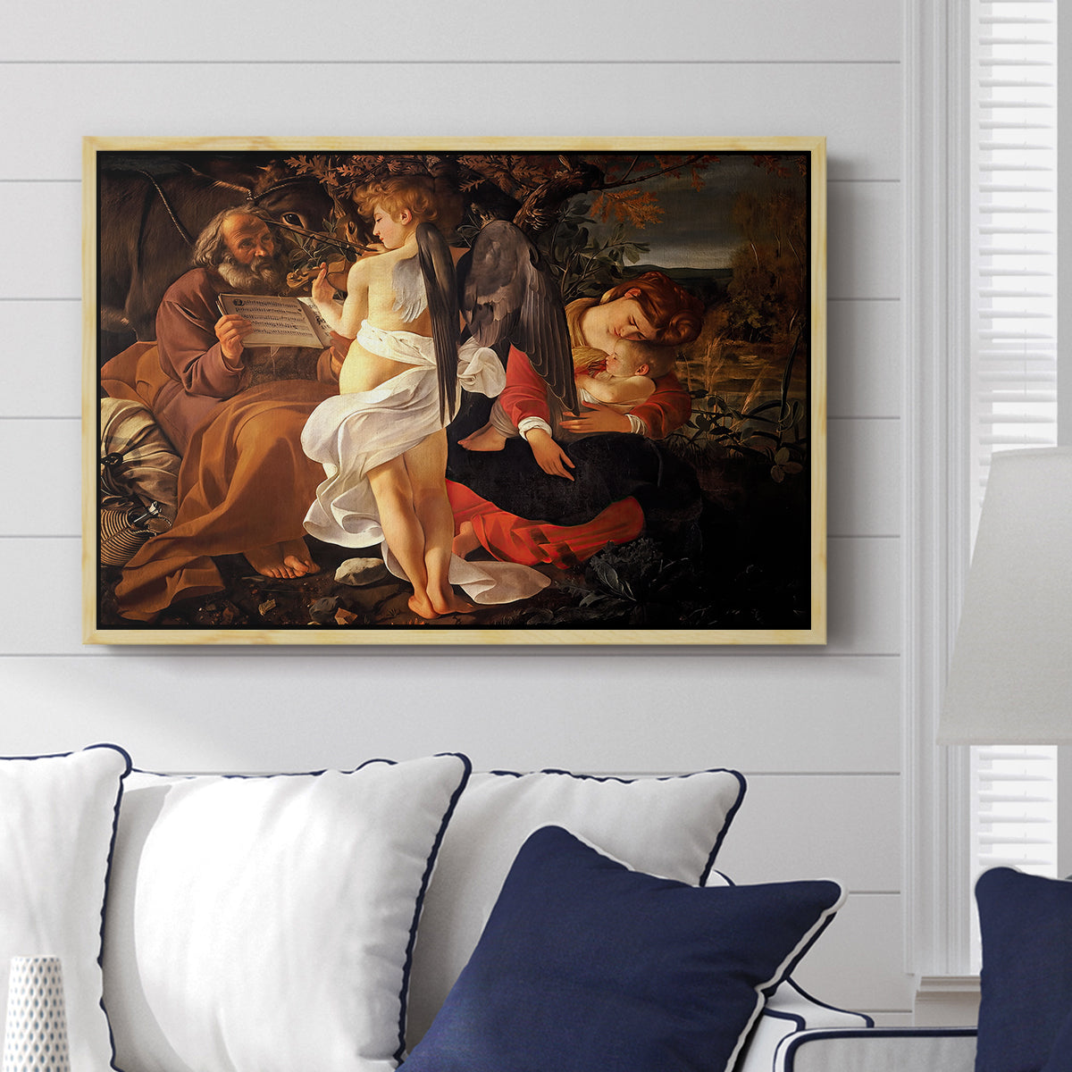 Rest on the Flight into Egypt, c. 1597 by Caravaggio displayed above a sofa in a living room in a natural wood floater frame, featuring a biblical scene of Mary, Joseph, and infant resting with angel musician.
