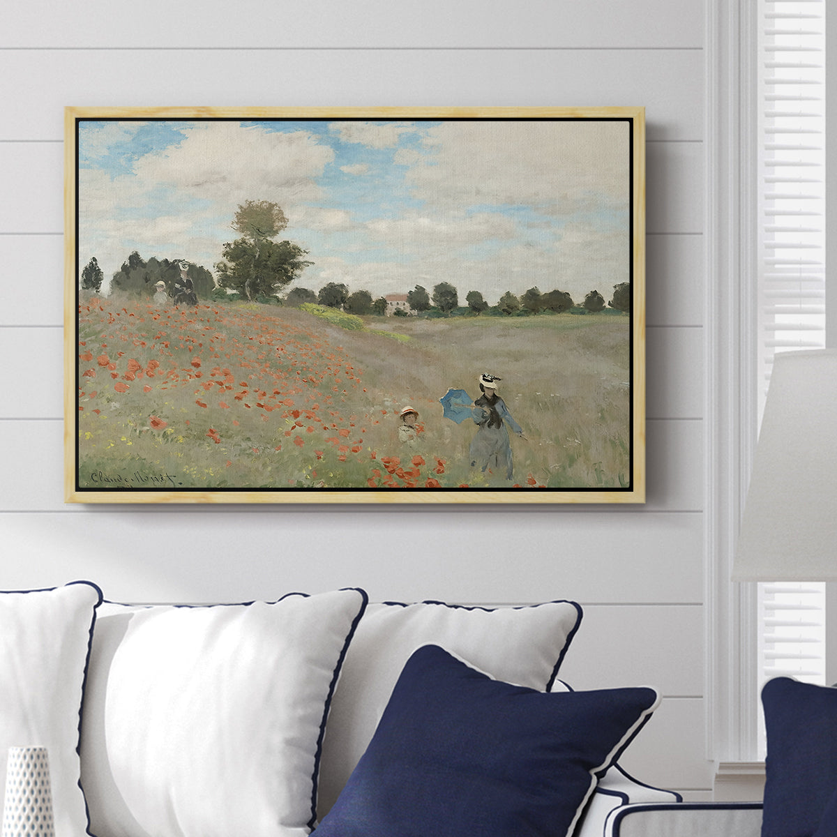 The Poppy Field near Argenteuil, 1873 by Claude Monet displayed above a sofa in a living room in a natural wood floater frame, featuring an impressionist countryside scene with flowers and strolling figures.