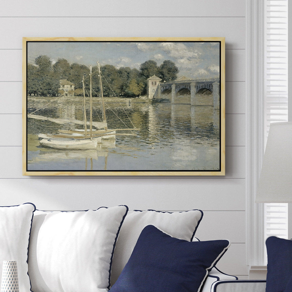 The Bridge at Argenteuil, 1874 by Claude Monet displayed above a sofa in a living room in a natural wood floater frame, featuring an impressionist river scene with boats passing beneath bridge.