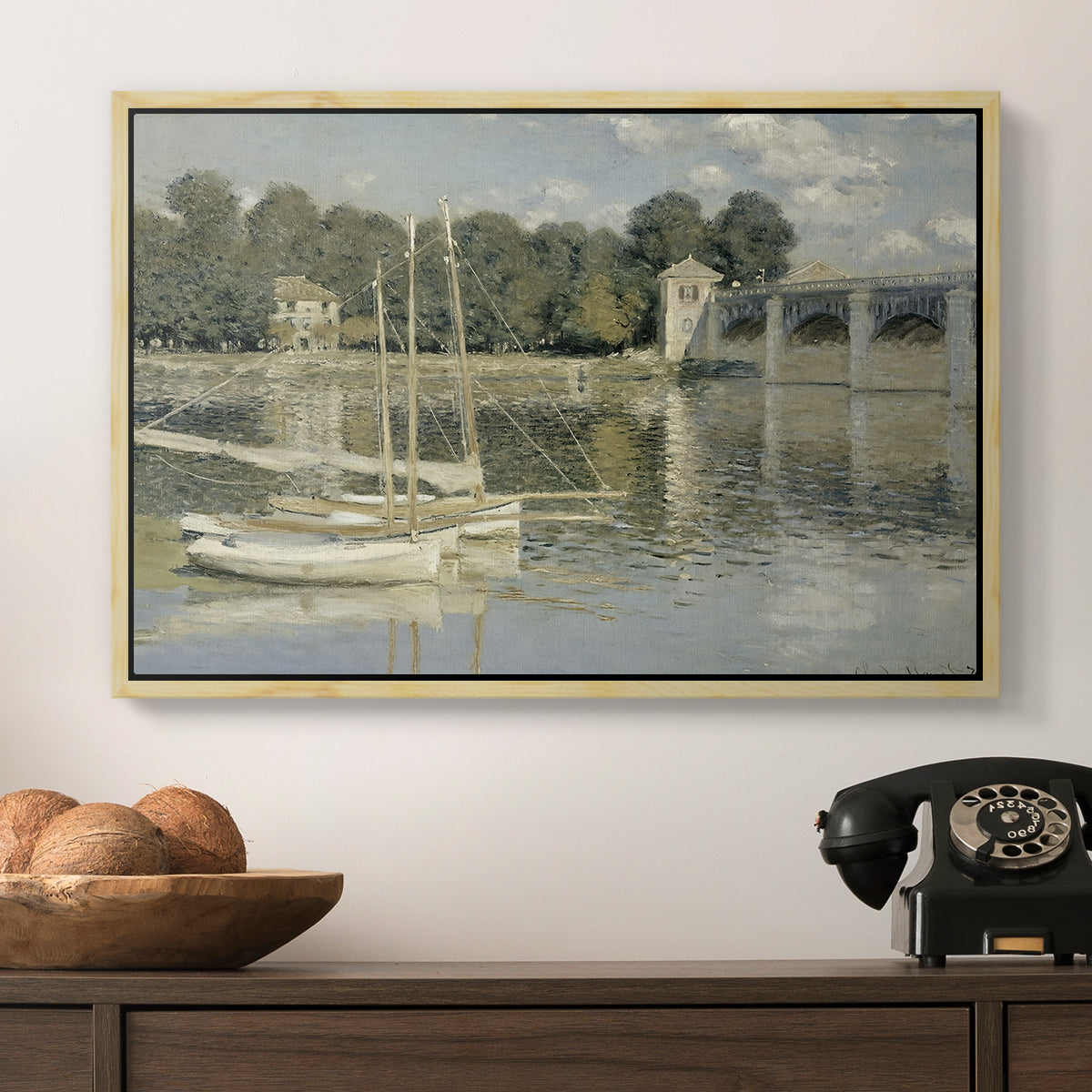 The Bridge at Argenteuil, 1874 by Claude Monet in a natural wood floater framed canvas displayed above a console table in a hallway, featuring a riverside view with architecture and nautical activity.