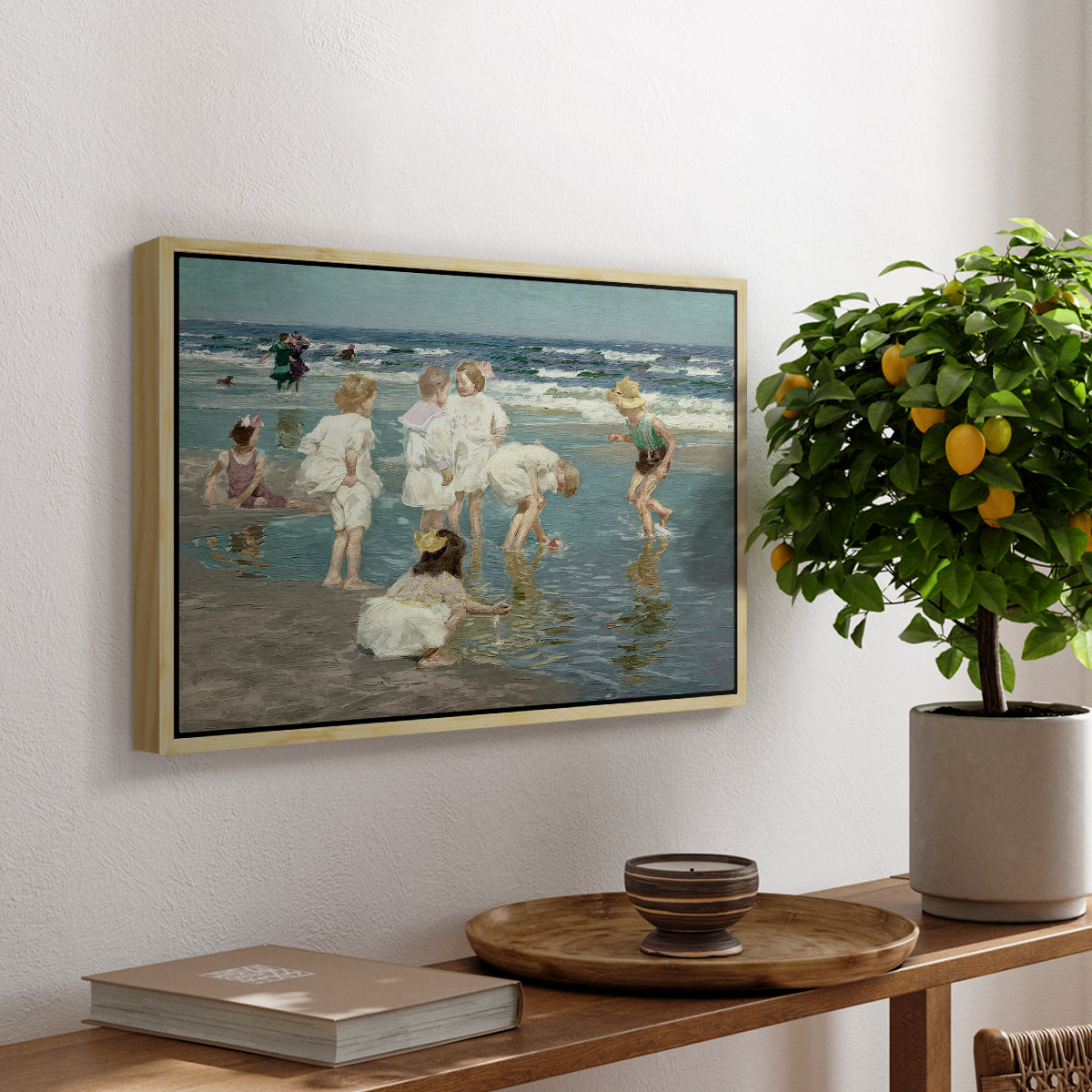 A Holiday, c. 1915 by Edward Henry Potthast, presented in a natural wood floater framed canvas featuring people relaxing and wading in shallow ocean water along a sunny beach, displayed above a hallway console table.
