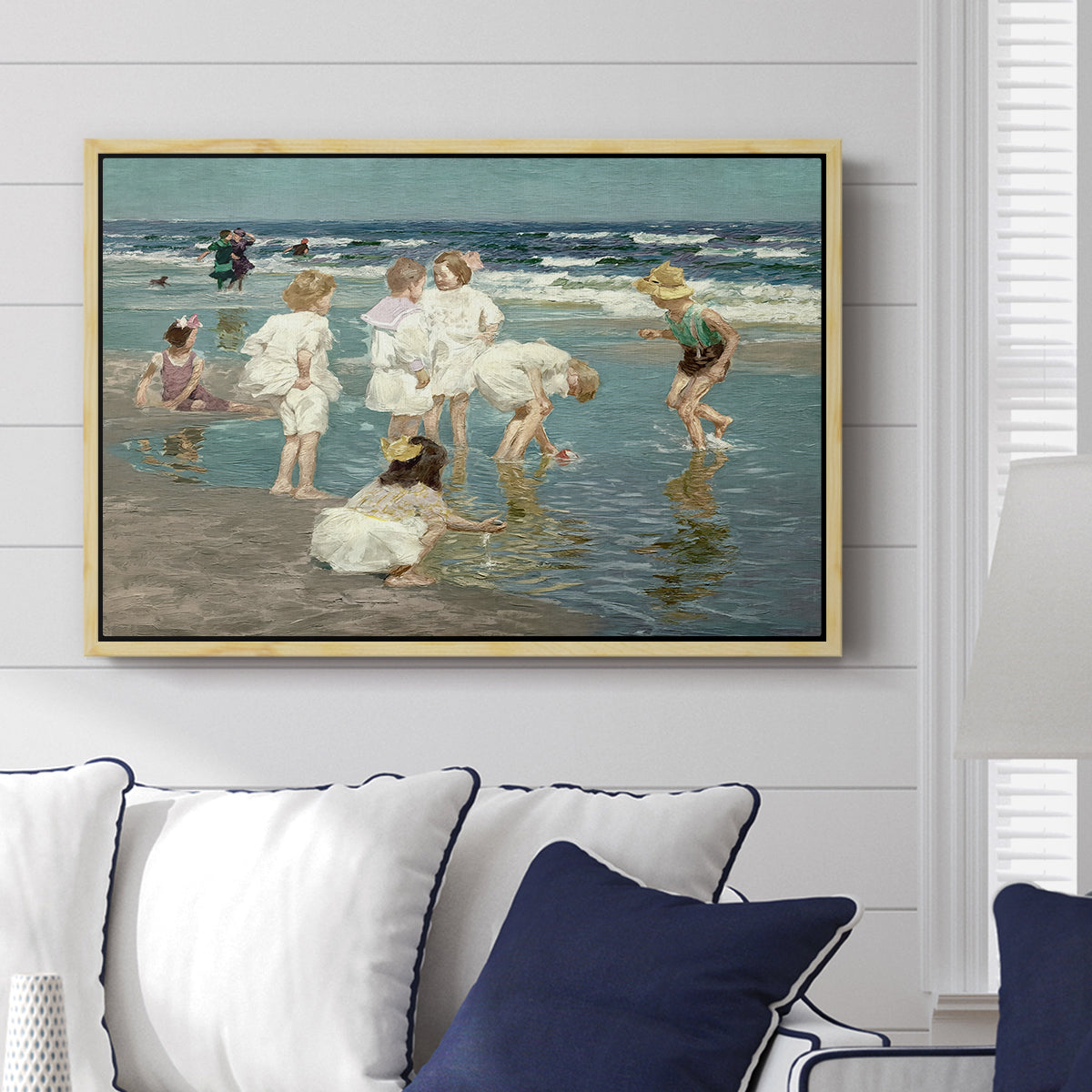 A Holiday, c. 1915 by Edward Henry Potthast displayed above a sofa in a living room in a natural wood floater frame, featuring a beach scene with figures bathing and standing in waves under bright sky.