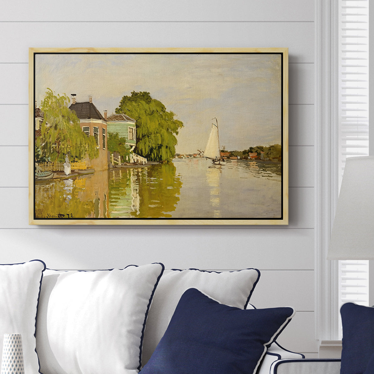 Houses on the Achterzaan, 1871 by Claude Monet displayed above a sofa in a living room in a natural wood floater frame, featuring riverside homes with trees and mirrored reflections in water.