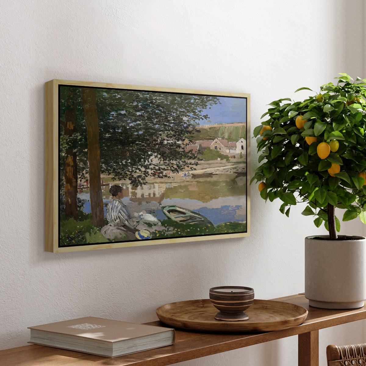 On the Bank of the Seine, Bennecourt, 1868 by Claude Monet, presented in a natural wood floater framed canvas featuring a woman and child seated along riverbank beneath trees in soft daylight, displayed above a hallway console table.
