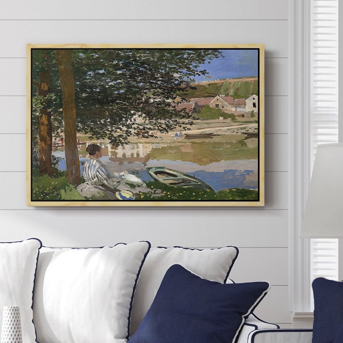 On the Bank of the Seine, Bennecourt, 1868 by Claude Monet displayed above a sofa in a living room in a natural wood floater frame, featuring a peaceful riverside scene with figures resting in grassy area by water.