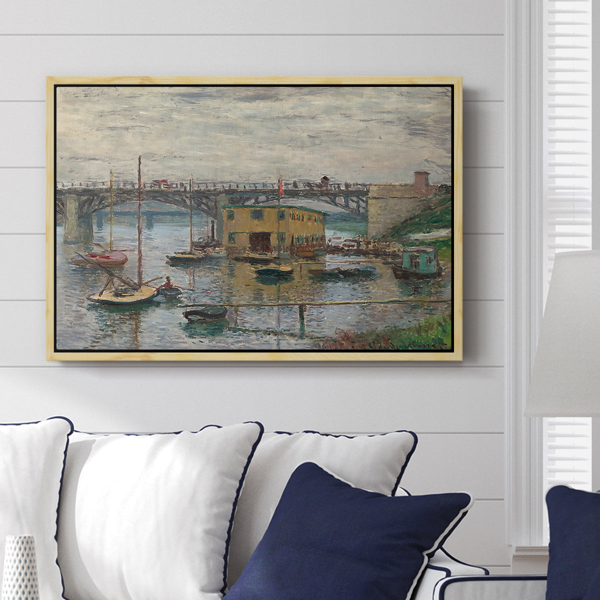 Bridge at Argenteuil on a Gray Day, c. 1876 by Claude Monet displayed above a sofa in a living room in a natural wood floater frame, featuring an impressionist riverscape with overcast atmosphere and soft tones.