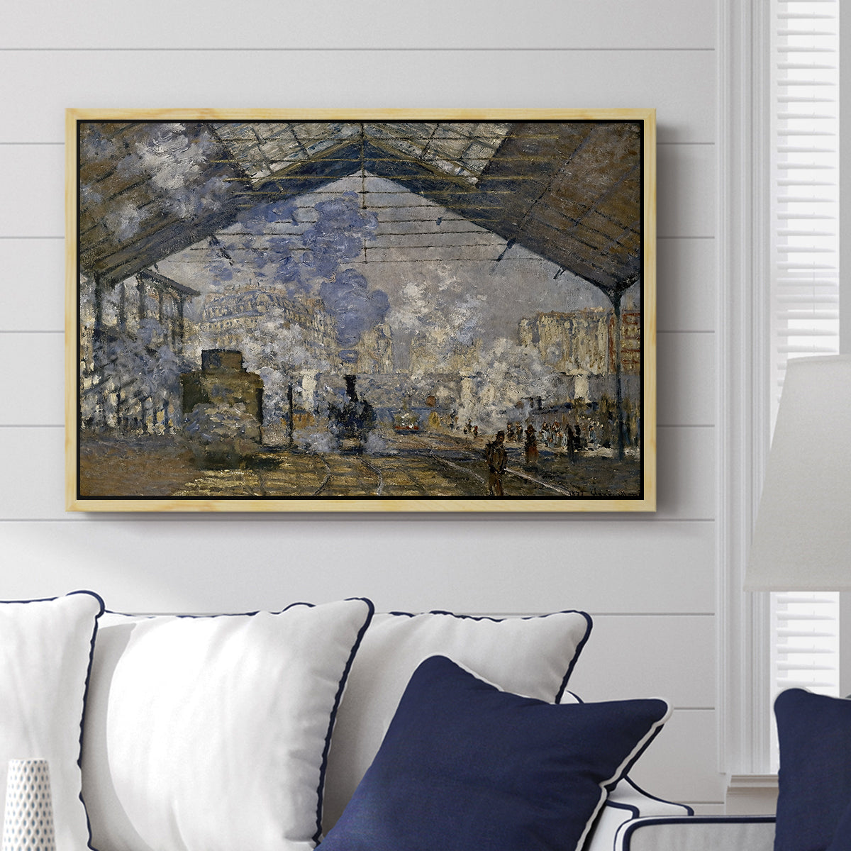 The Gare Saint-Lazare, 1877 by Claude Monet displayed above a sofa in a living room in a natural wood floater frame, featuring an impressionist scene of trains and smoke inside busy railway station.