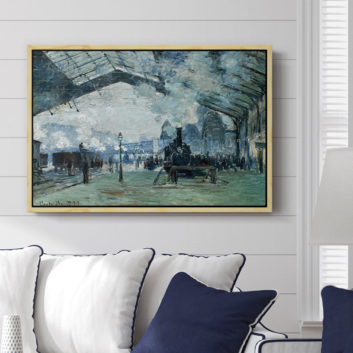 The Saint-Lazare Station, 1877 by Claude Monet displayed above a sofa in a living room in a natural wood floater frame, featuring an impressionist railway scene with locomotives and haze.