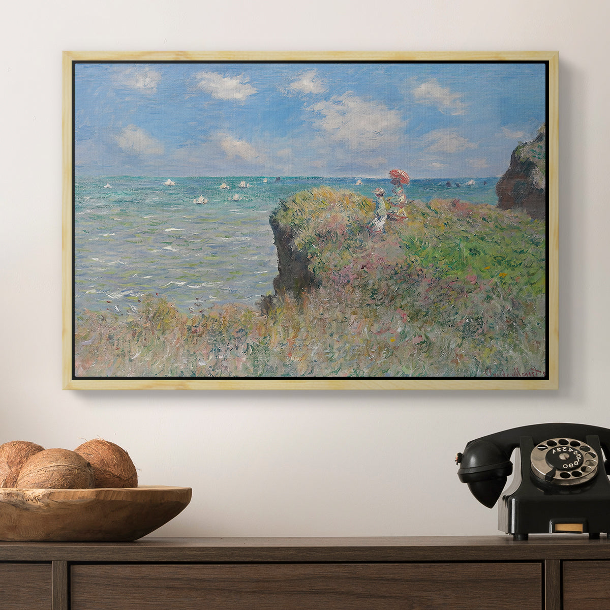 The Cliff Walk at Pourville, 1882 by Claude Monet in a natural wood floater framed canvas displayed above a console table in a hallway, featuring an elevated shoreline path with figures and bright sky.