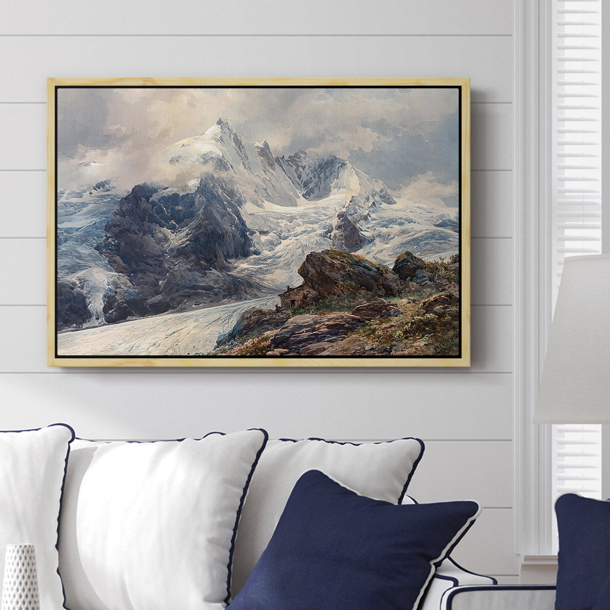The Grossglockner, 1880 by Edward Theodore Compton displayed above a sofa in a living room in a natural wood floater frame, featuring a dramatic mountain summit with snow and rugged slopes.