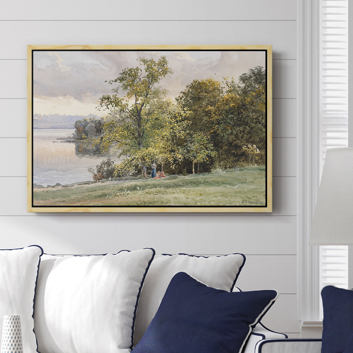 Landscape at Lake Starnberg, 1890 by Edward Theodore Compton displayed above a sofa in a living room in a natural wood floater frame, featuring a peaceful lake scene with reflections and surrounding greenery.