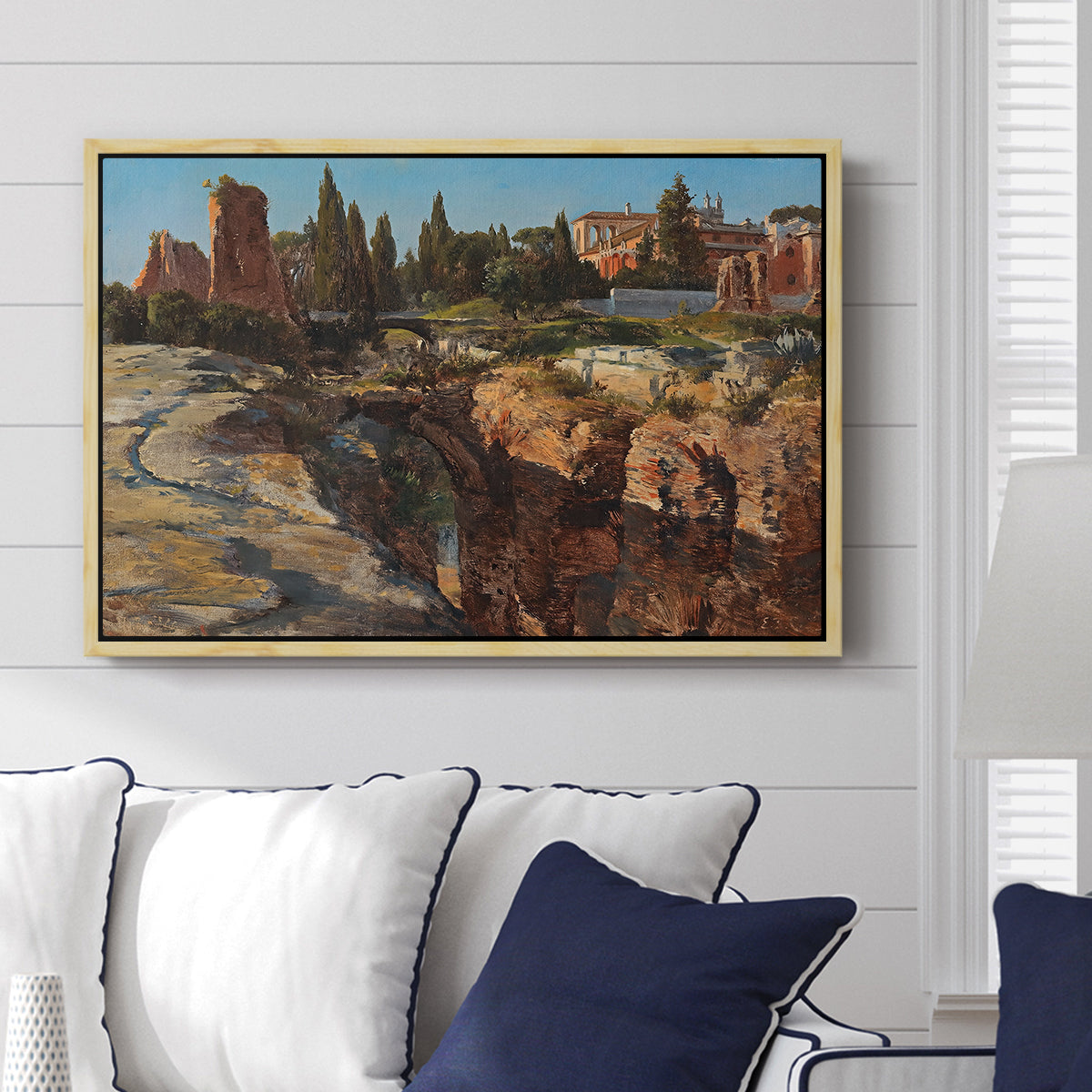 Palatine in Rome, c. 1921 by Edward Theodore Compton displayed above a sofa in a living room in a natural wood floater frame, featuring an architectural scene of ancient structures in sunlit environment.