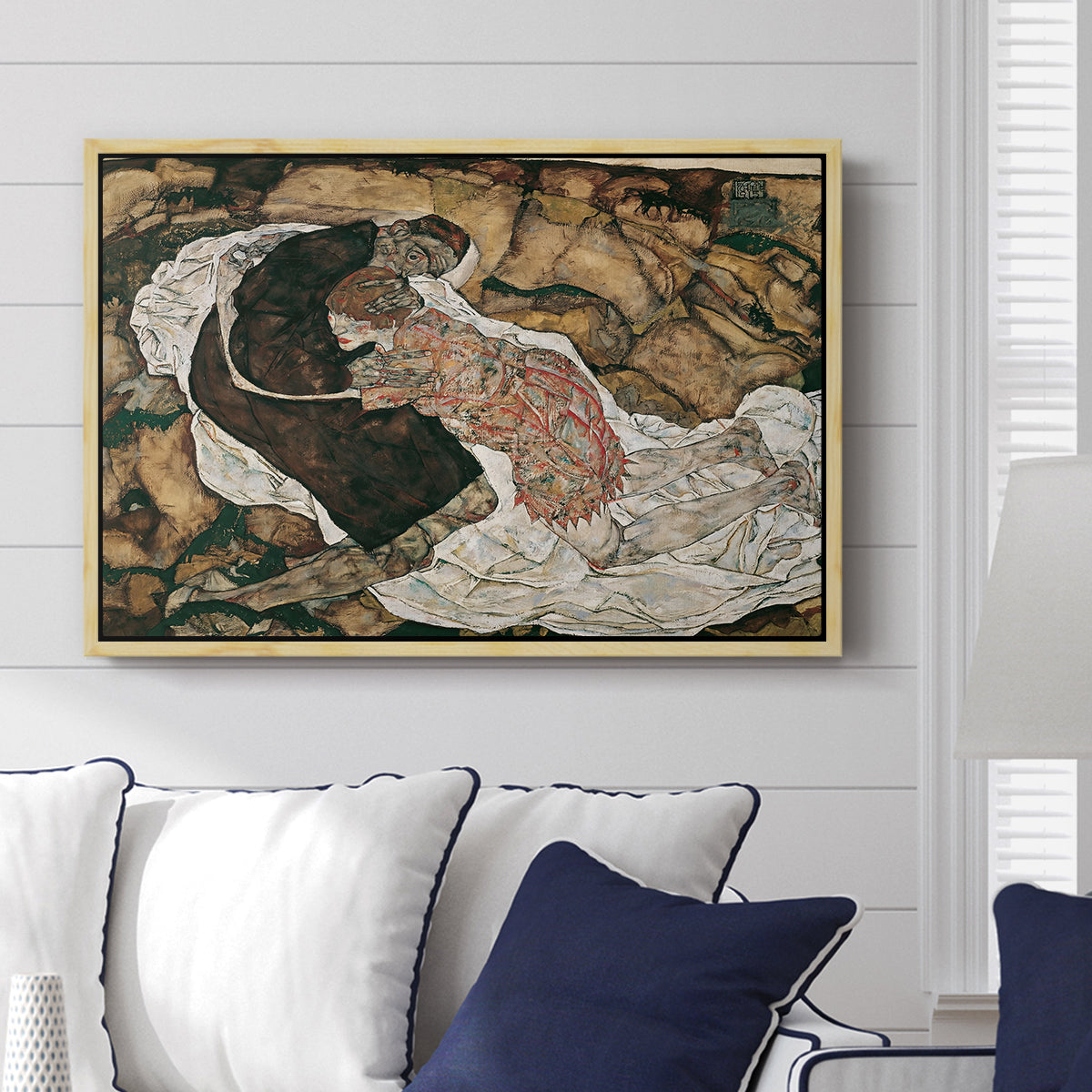 Death and the Maiden, 1915 by Egon Schiele displayed above a sofa in a living room in a natural wood floater frame, featuring a symbolic scene of mortality with two figures clinging together.