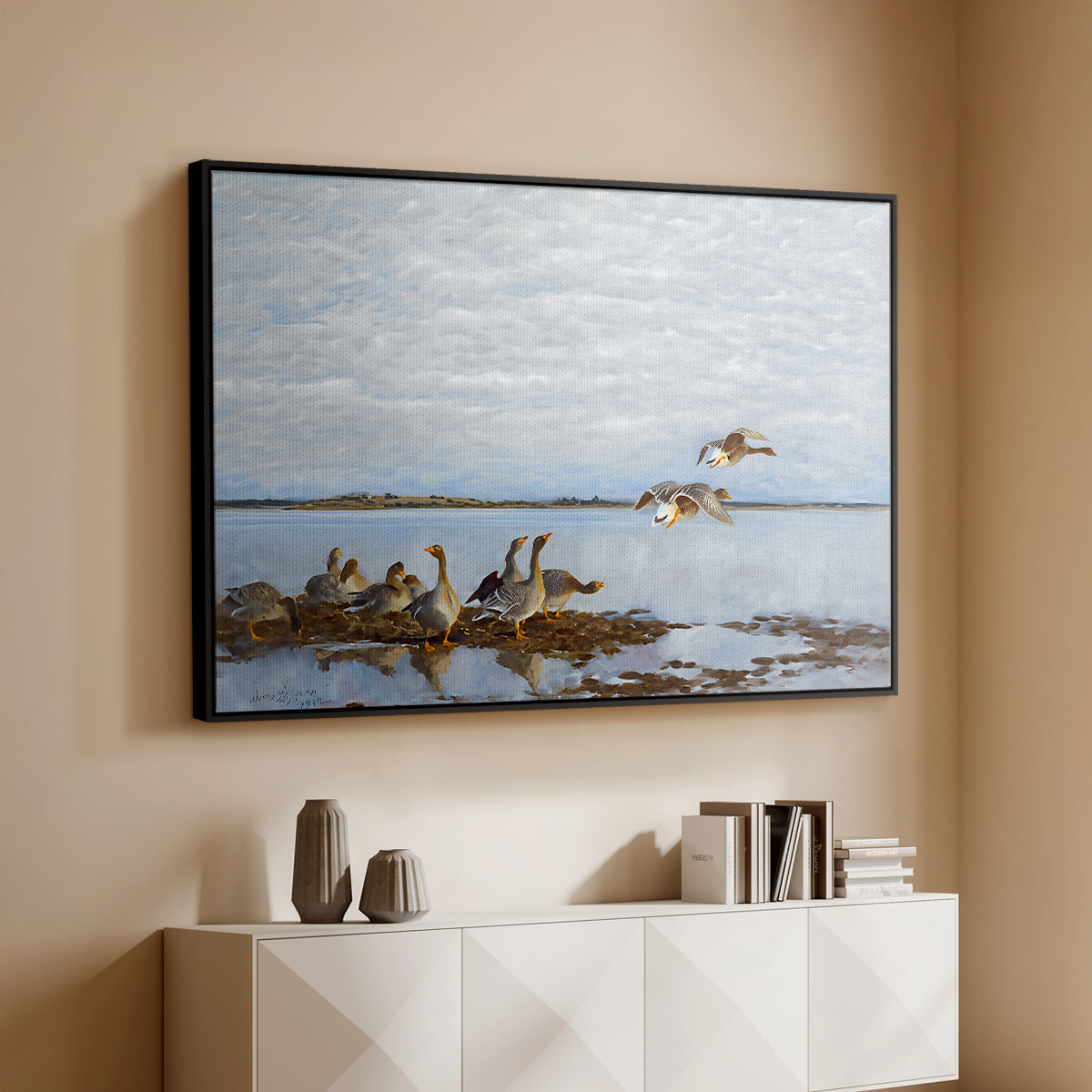 Black floater framed canvas of Greylag Geese (Grågäss), 1922 by Bruno Liljefors, featuring birds standing and resting along shoreline with soft grass and water above a console cabinet in a minimalist entryway.