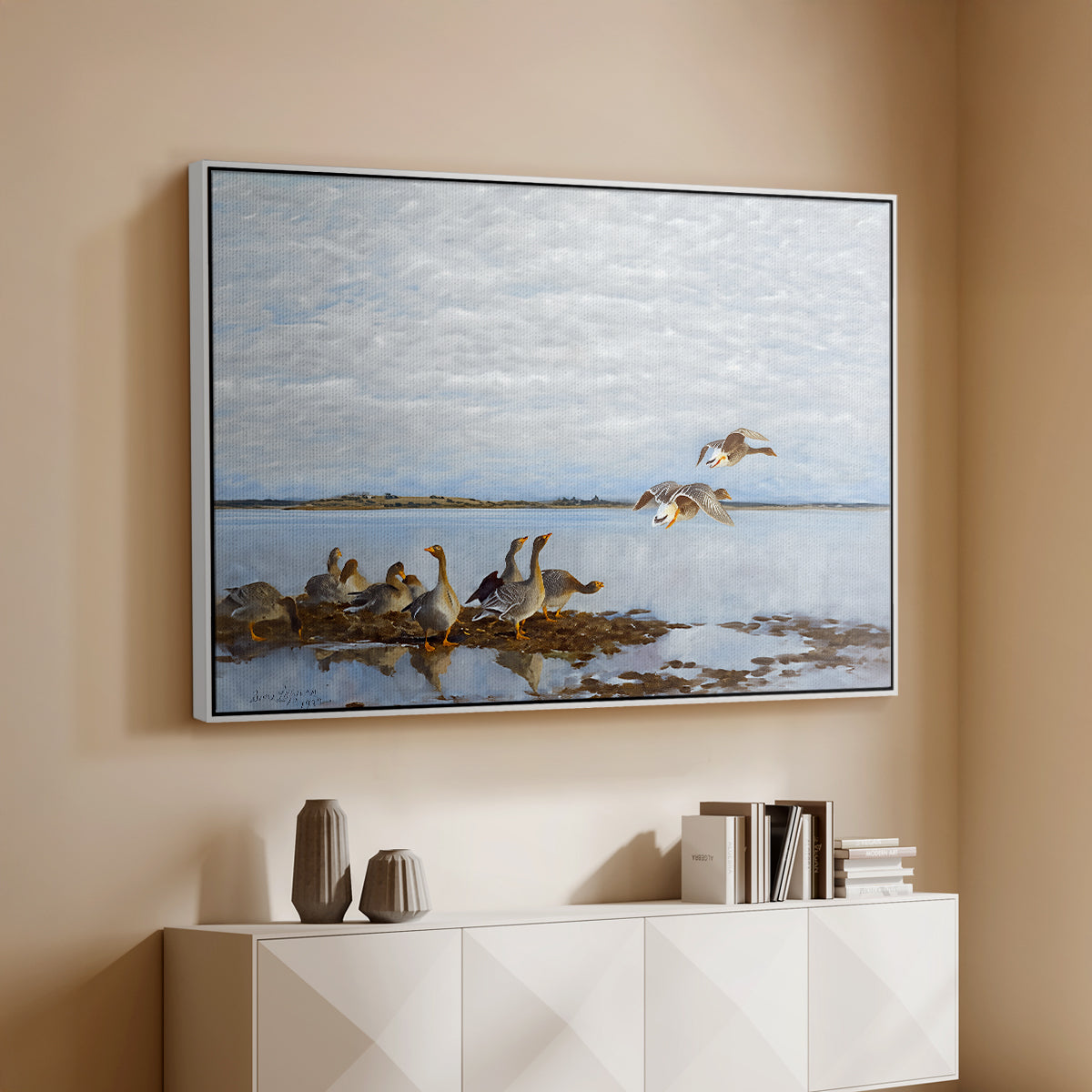 White floater framed canvas of Greylag Geese (Grågäss), 1922 by Bruno Liljefors, featuring birds standing and resting along shoreline with soft grass and water above a console cabinet in a minimalist entryway.
