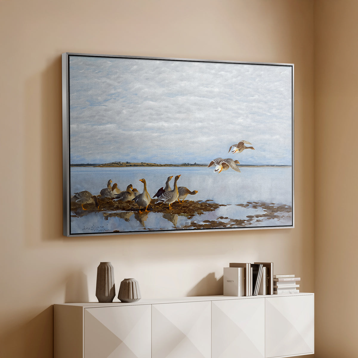 Silver floater framed canvas of Greylag Geese (Grågäss), 1922 by Bruno Liljefors, featuring birds standing and resting along shoreline with soft grass and water above a console cabinet in a minimalist entryway.