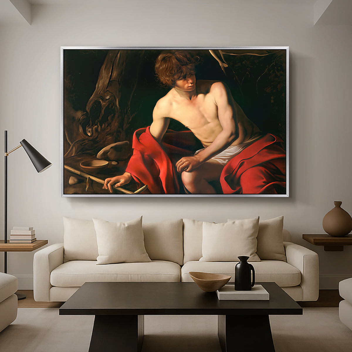 Saint John the Baptist in the Wilderness, c. 1603-1604 by Caravaggio as a silver floater framed canvas featuring a young Saint John seated in wilderness with staff and red drapery, displayed above a sofa in a modern living room.