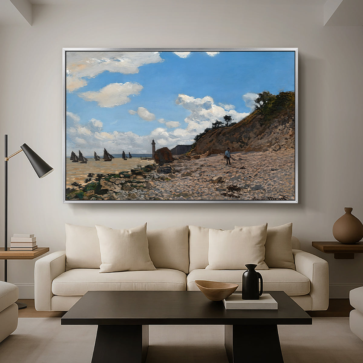 The Beach at Honfleur, 1864 by Claude Monet as a silver floater framed canvas featuring a coastal beach scene with figures gathered along shoreline and boats nearby, displayed above a sofa in a modern living room.