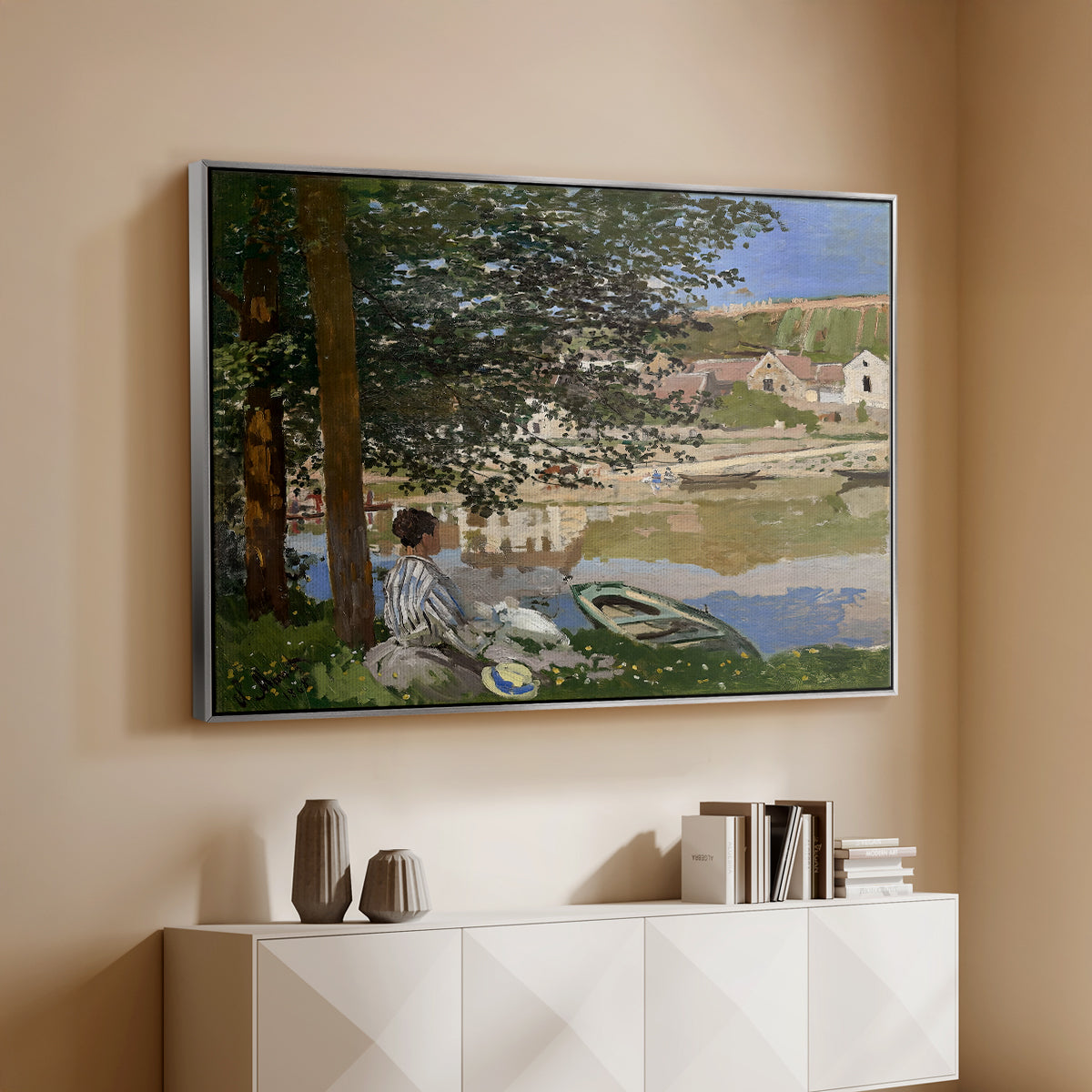 Silver floater framed canvas of On the Bank of the Seine, Bennecourt, 1868 by Claude Monet, featuring a peaceful riverside scene with figures resting in grassy area by water above a console cabinet in a minimalist entryway.