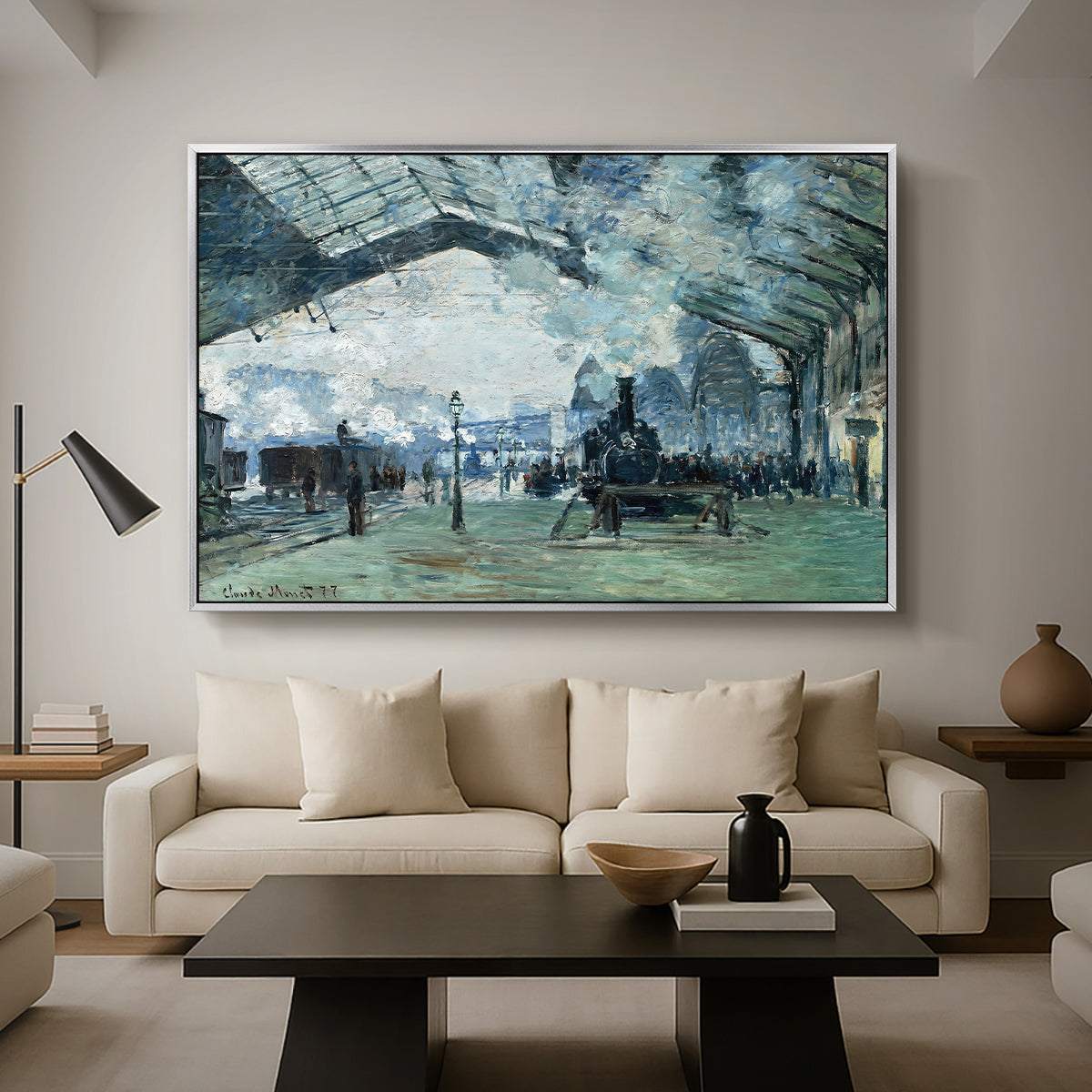 The Saint-Lazare Station, 1877 by Claude Monet as a silver floater framed canvas featuring trains arriving under large arched station roof with billowing steam, displayed above a sofa in a modern living room.