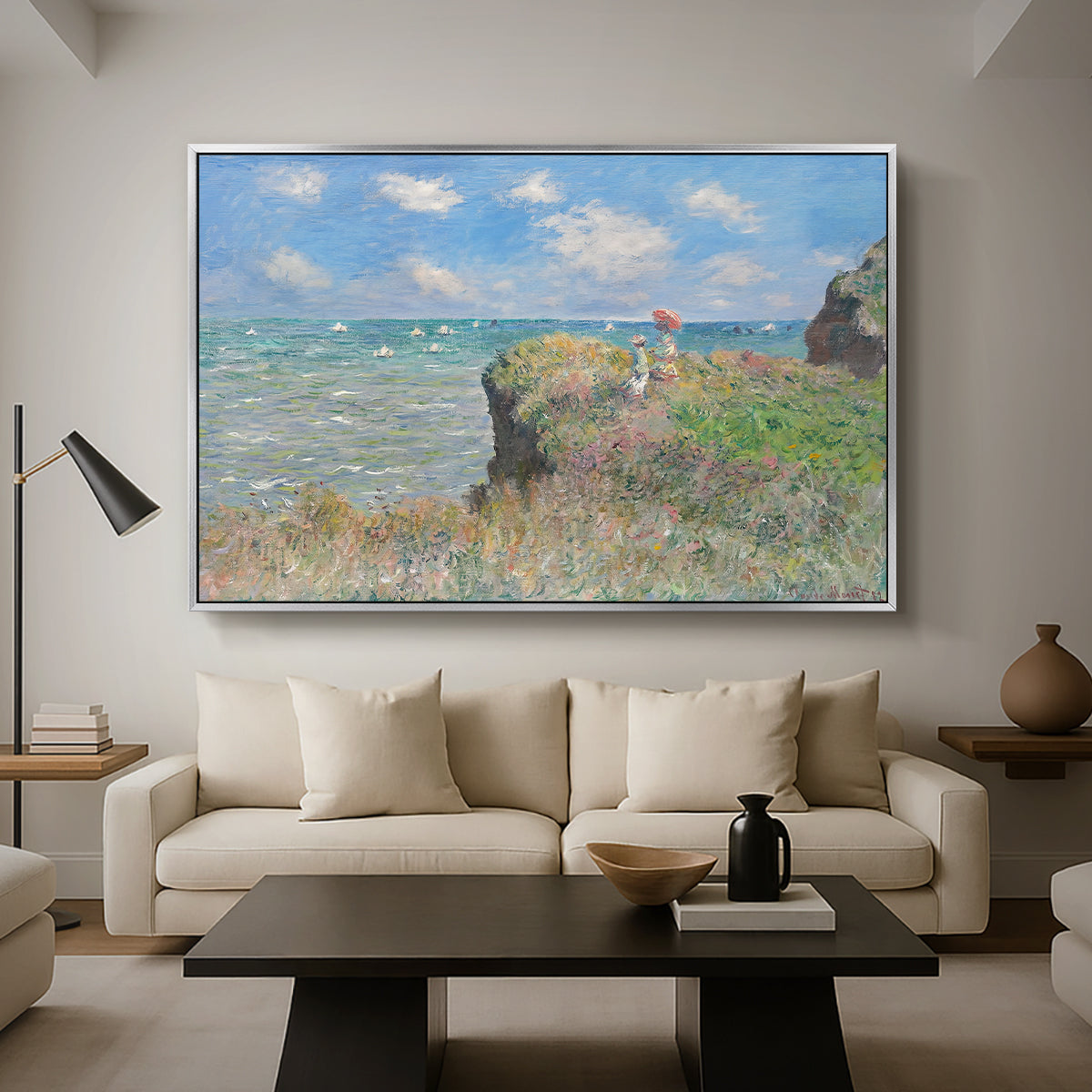 The Cliff Walk at Pourville, 1882 by Claude Monet as a silver floater framed canvas featuring figures walking along coastal cliff path overlooking ocean and sky, displayed above a sofa in a modern living room.