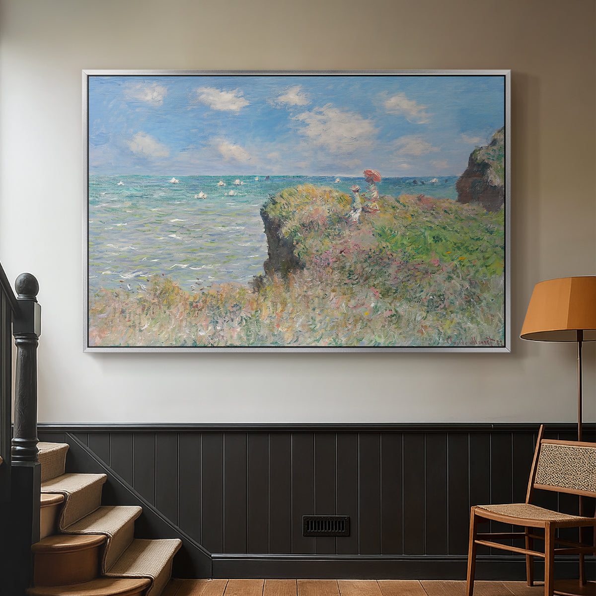Silver floater framed canvas featuring an elevated shoreline path with figures and bright sky, displayed in a hallway near a staircase.